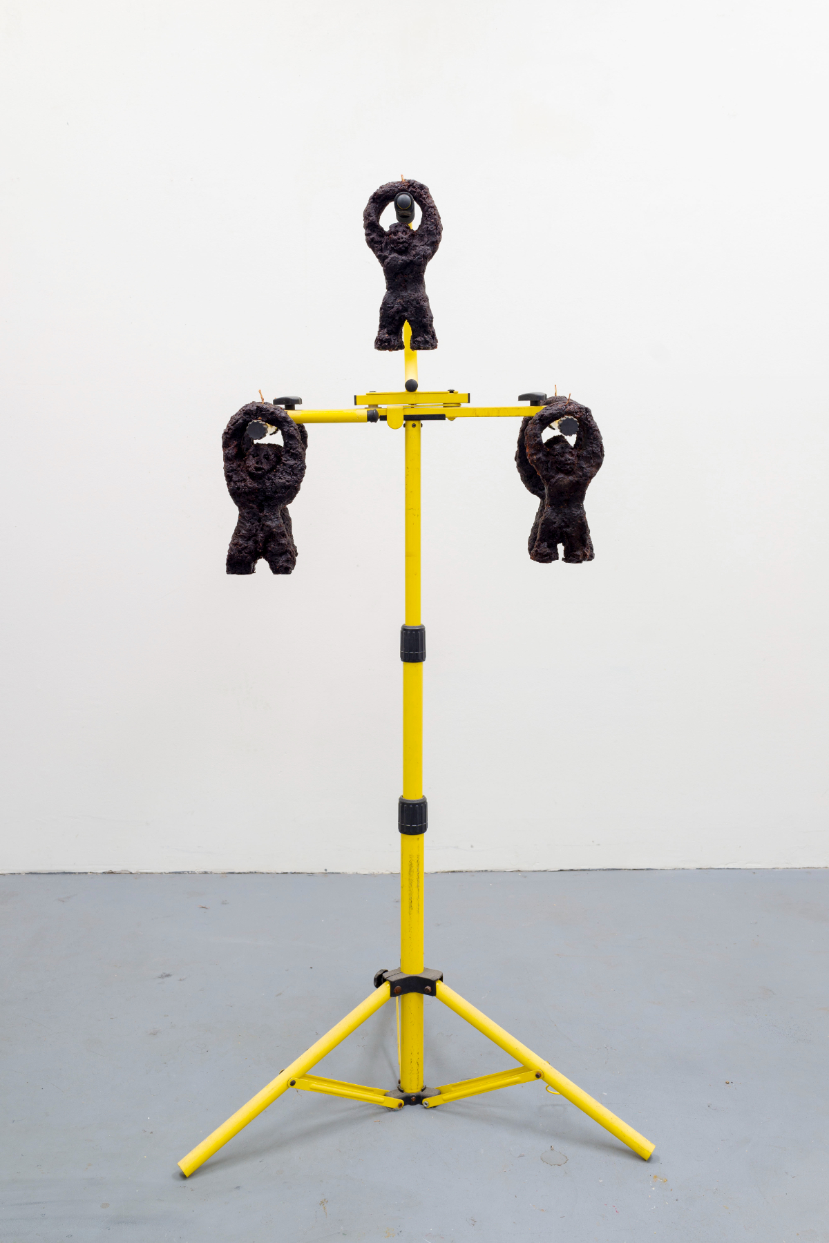 Candelabra 2 - A Sculpture by Brian Dario, A Worksite light-stand with five dark wax ape-like figures with wicks pointing up from their hands hanging from different points on the light-stand.  One figure hangs from a higher point, while the other four are symmetrically balanced in groups of two on either side.  The front two figures obscure the back two figures from this angle, making it look as though there are only three figures