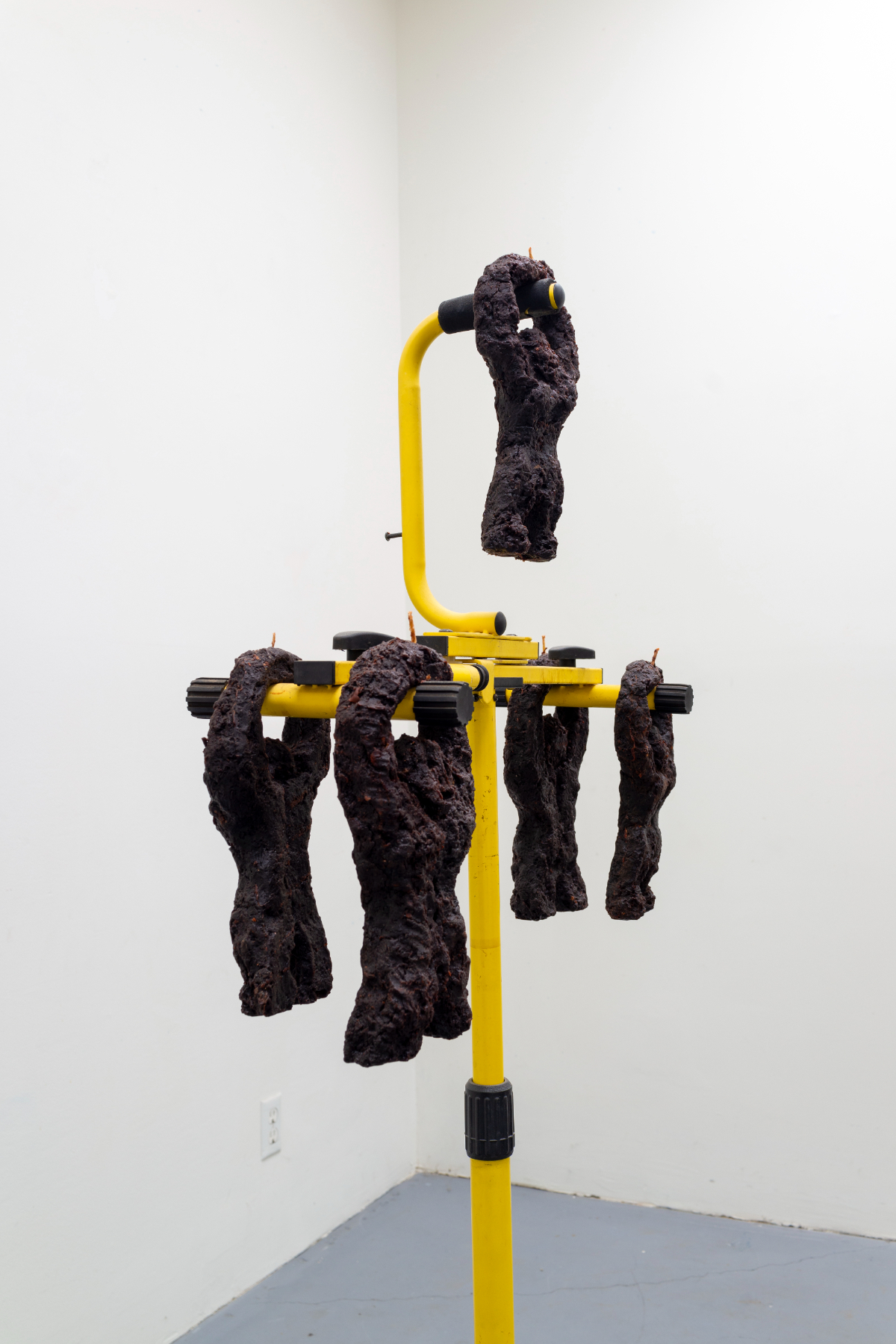 Candelabra 2 - A Sculpture by Brian Dario, A side view of the top of a worksite light-stand with five dark wax ape like figures with wicks pointing up from their hands hanging from different points on the light stand.  One figure hangs from a higher point, while the other four are symmetrically balanced in groups of two on either side.  All five figures are visible from this angle, as well as their wicks