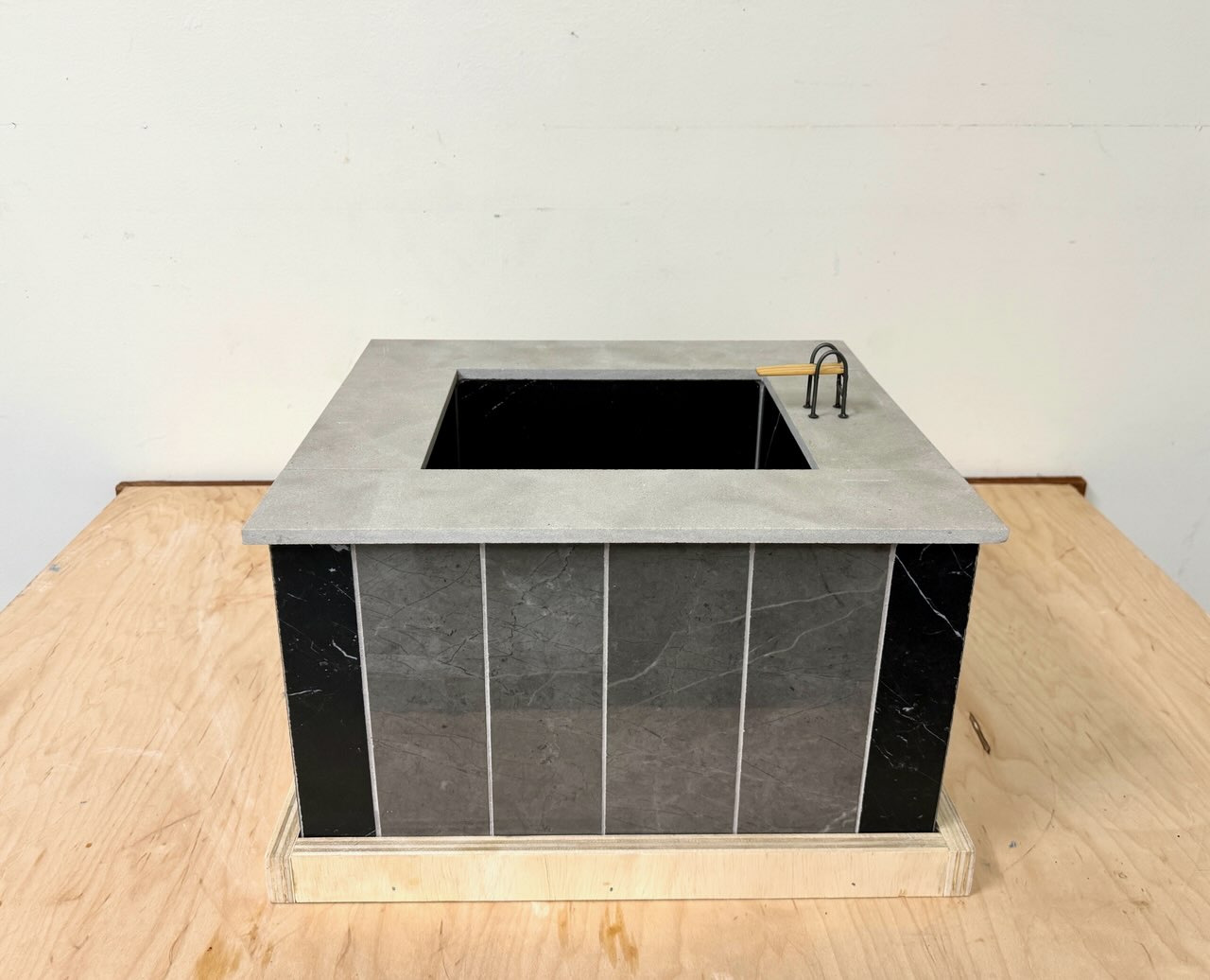 Swimming Pool Monument (for Keith) - A Sculpture by Brian Dario, A small rectangular swimming pool containing no water, with a small diving board on the lip, a light gray lip around the edge of the pool, walls consisting of tiles of different colors: one dark almost black tile in each corner, four medium gray tiles in the center of each wall, white grout separating the tiles, a dark almost black color on the inside of the pool, resting on a wooden table against a white wall.