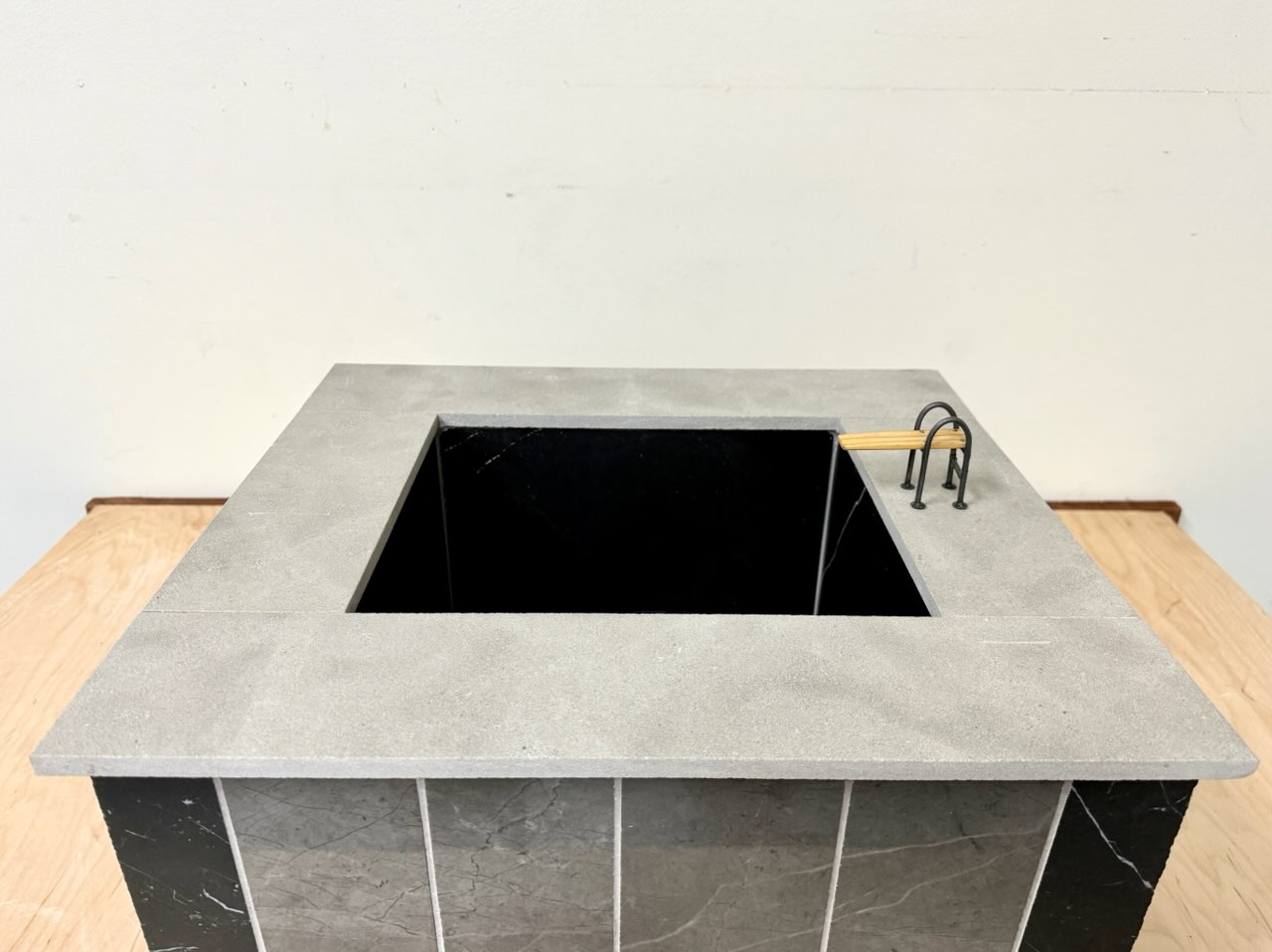 Swimming Pool Monument (for Keith) - A Sculpture by Brian Dario, A small rectangular swimming pool containing no water, with a small diving board on the lip, a light gray lip around the edge of the pool, walls consisting of tiles of different colors: one dark almost black tile in each corner, four medium gray tiles in the center of each wall, white grout separating the tiles, a dark almost black color on the inside of the pool, resting on a wooden table against a white wall. At a higher angle