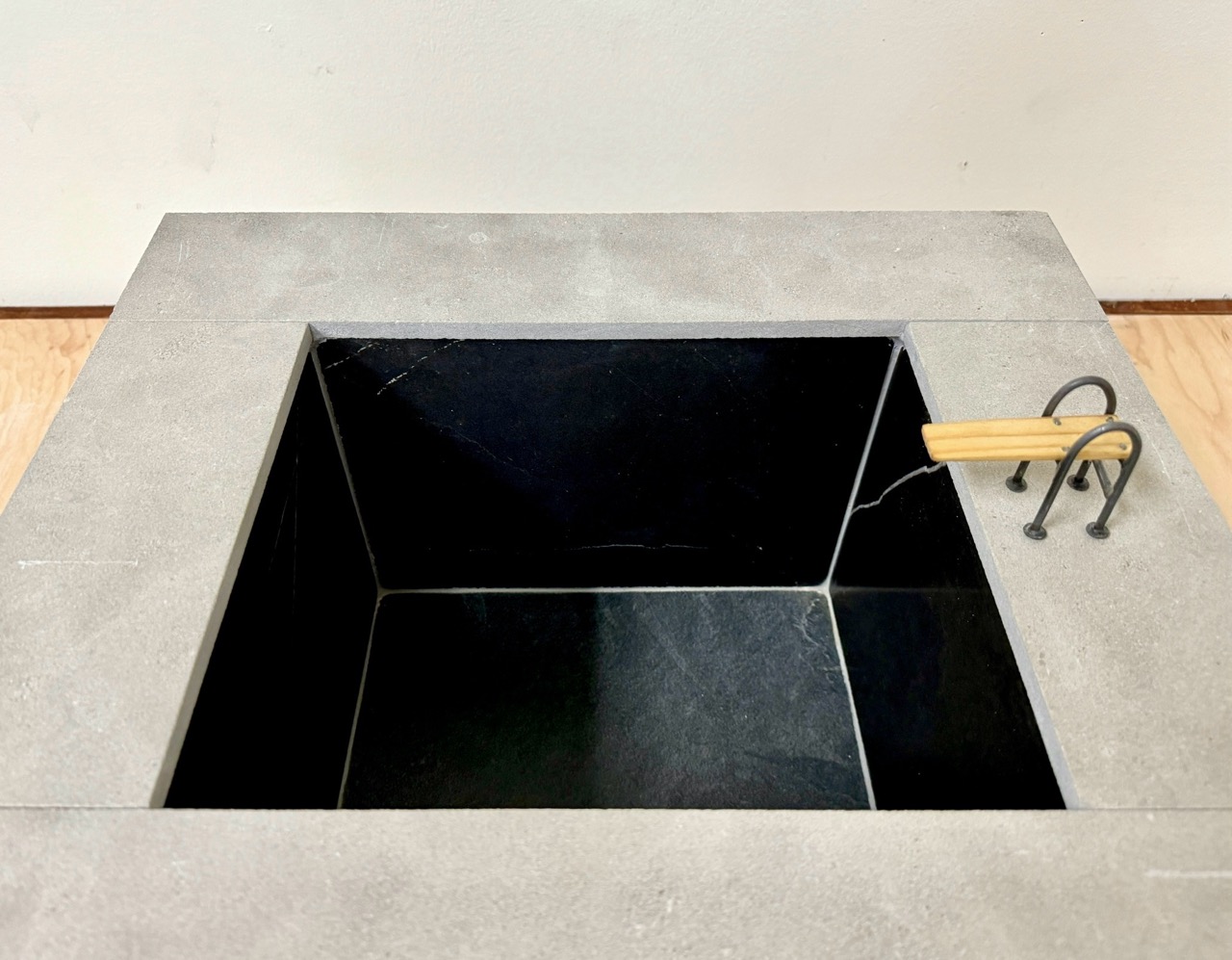 Swimming Pool Monument (for Keith) - A Sculpture by Brian Dario, A small rectangular swimming pool containing no water, with a small diving board on the lip, a light gray lip around the edge of the pool, walls consisting of tiles of different colors: one dark almost black tile in each corner, four medium gray tiles in the center of each wall, white grout separating the tiles, a dark almost black color on the inside of the pool, resting on a wooden table against a white wall. At an even higher angle, the detail on the diving board is now visible, the inside of the pool's white grout is now visible.